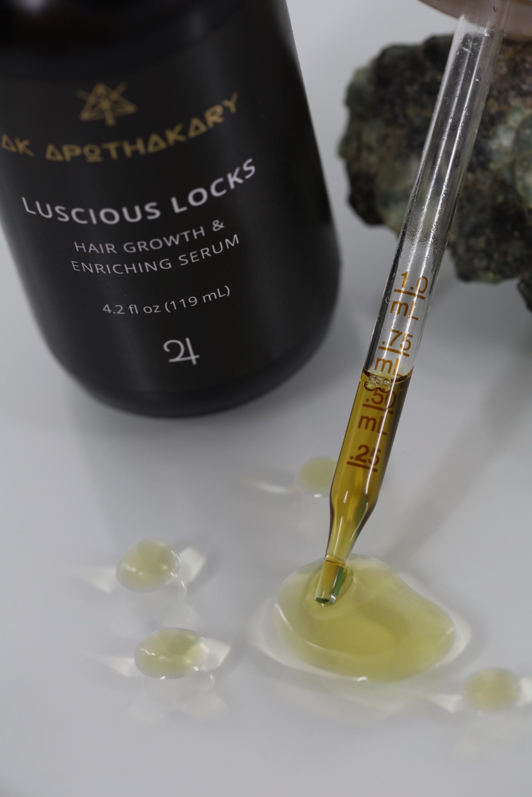 An eye dropper drips the Luscious Locks serum on glass in front of a bottle of the product.